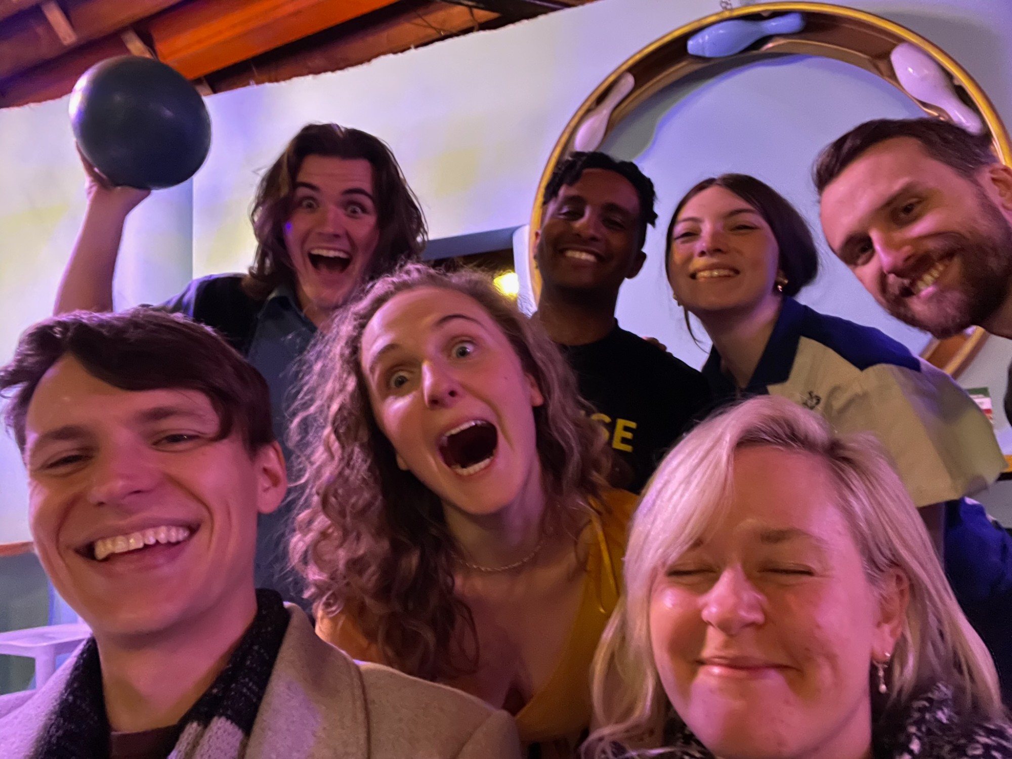 Hope shouting with delight in a selfie with six friends at a bowling alley, one holding a ball aloft