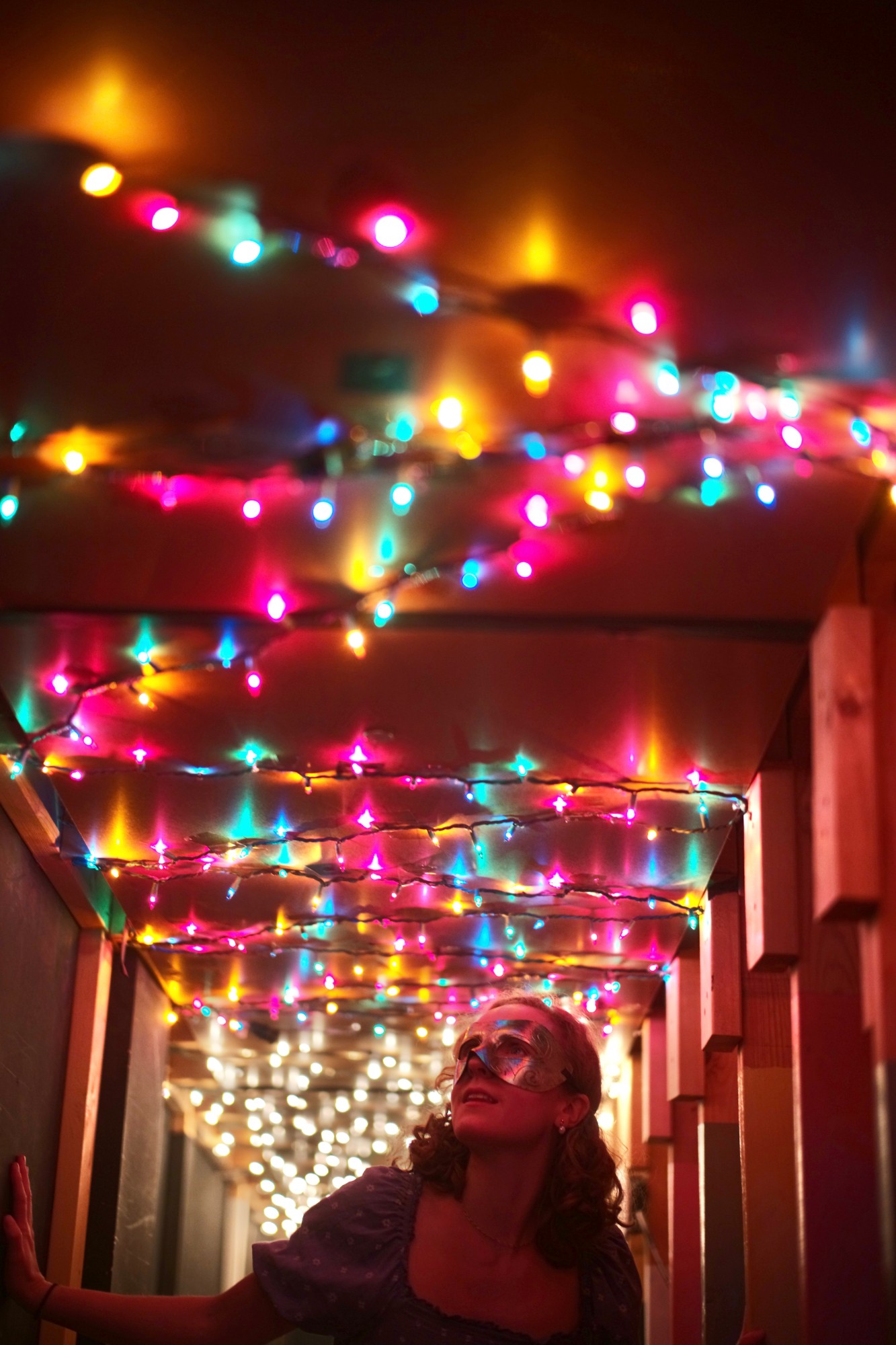 Hope in a masquerade mask gazing up at a corridor ceiling strung with colourful fairy lights