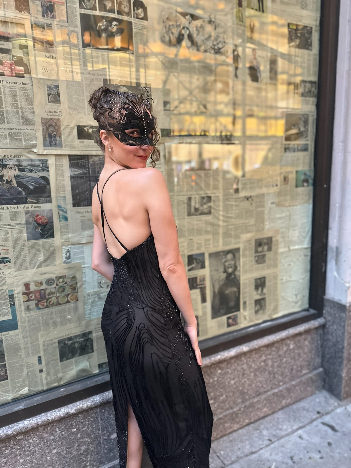 Hope in a lace masquerade mask and black beaded gown, glancing back over her shoulder in front of a newspaper-covered window