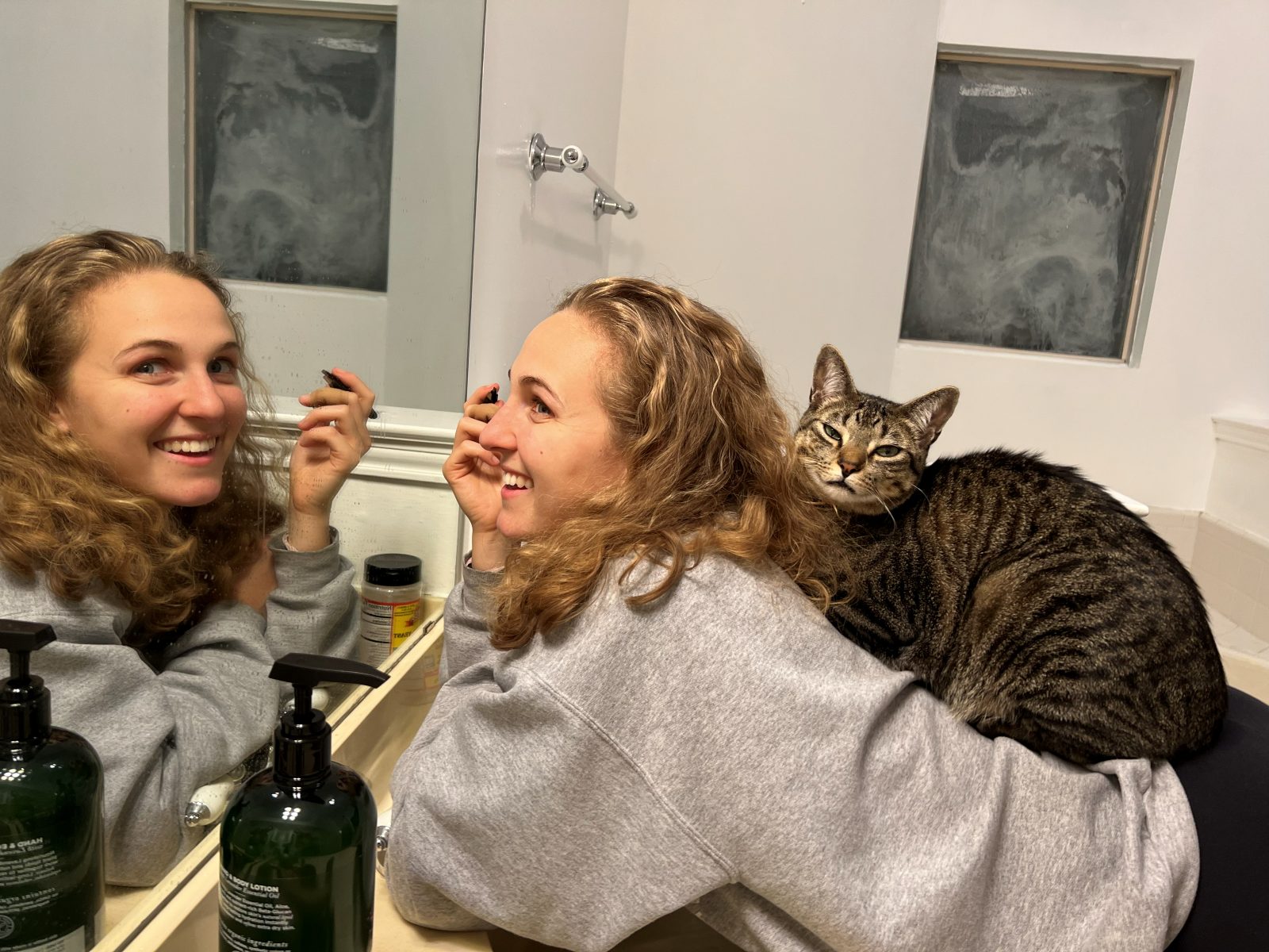 Hope laughing at a bathroom mirror while a tabby cat perches across her back