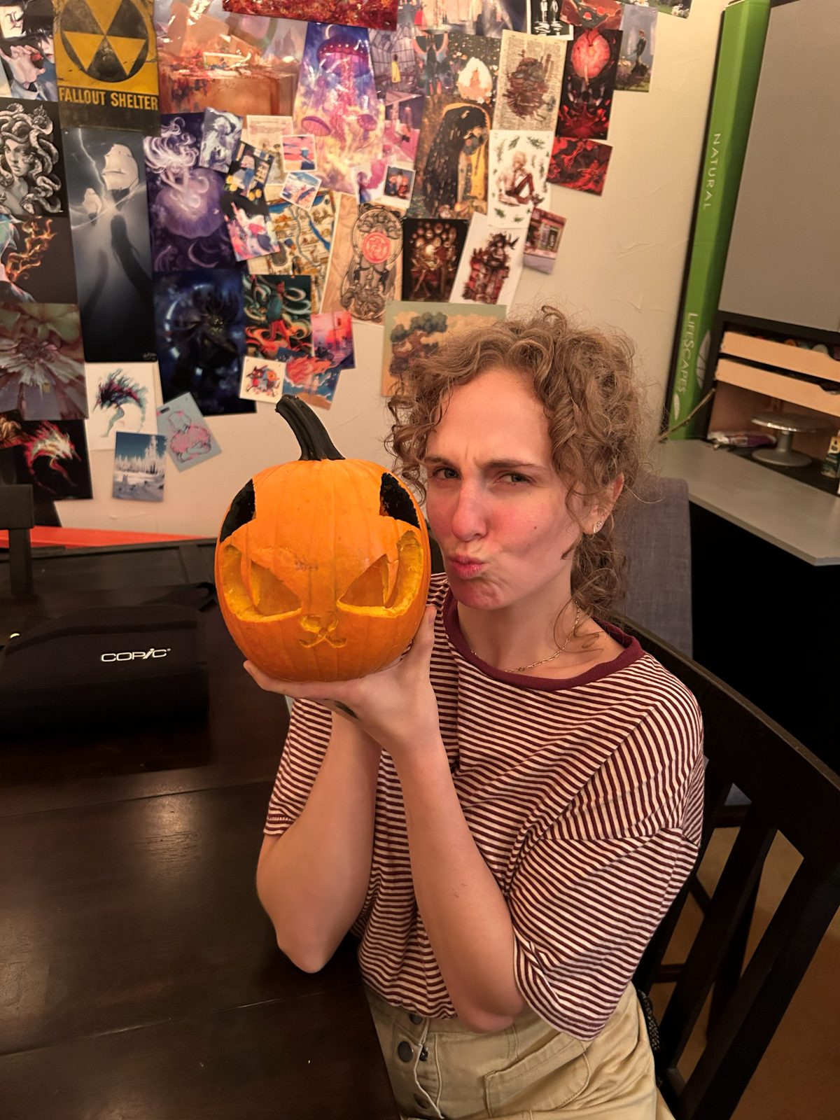 Hope pulling a face while holding up a cat-shaped carved pumpkin, a wall of art prints behind her