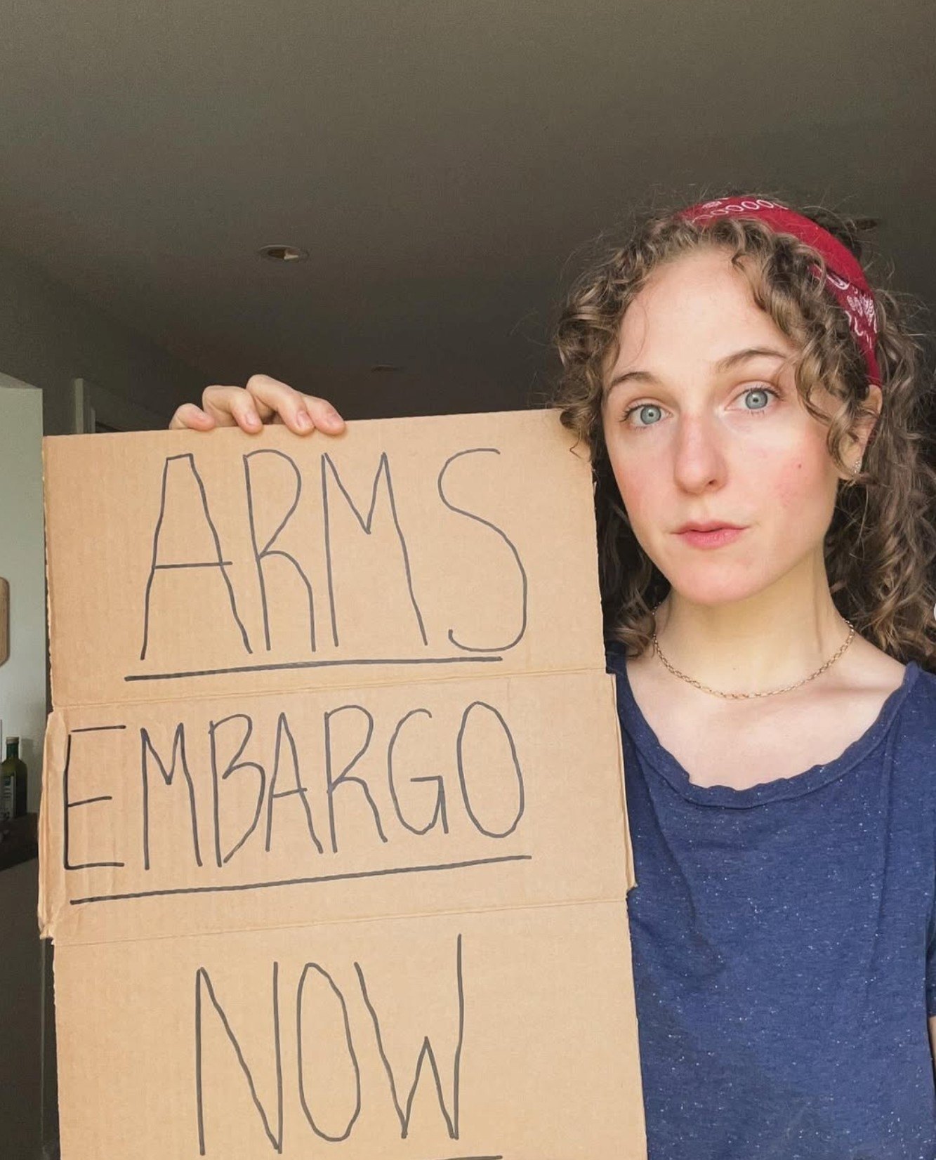 Hope in a red bandana holding a hand-lettered cardboard sign reading Arms Embargo Now