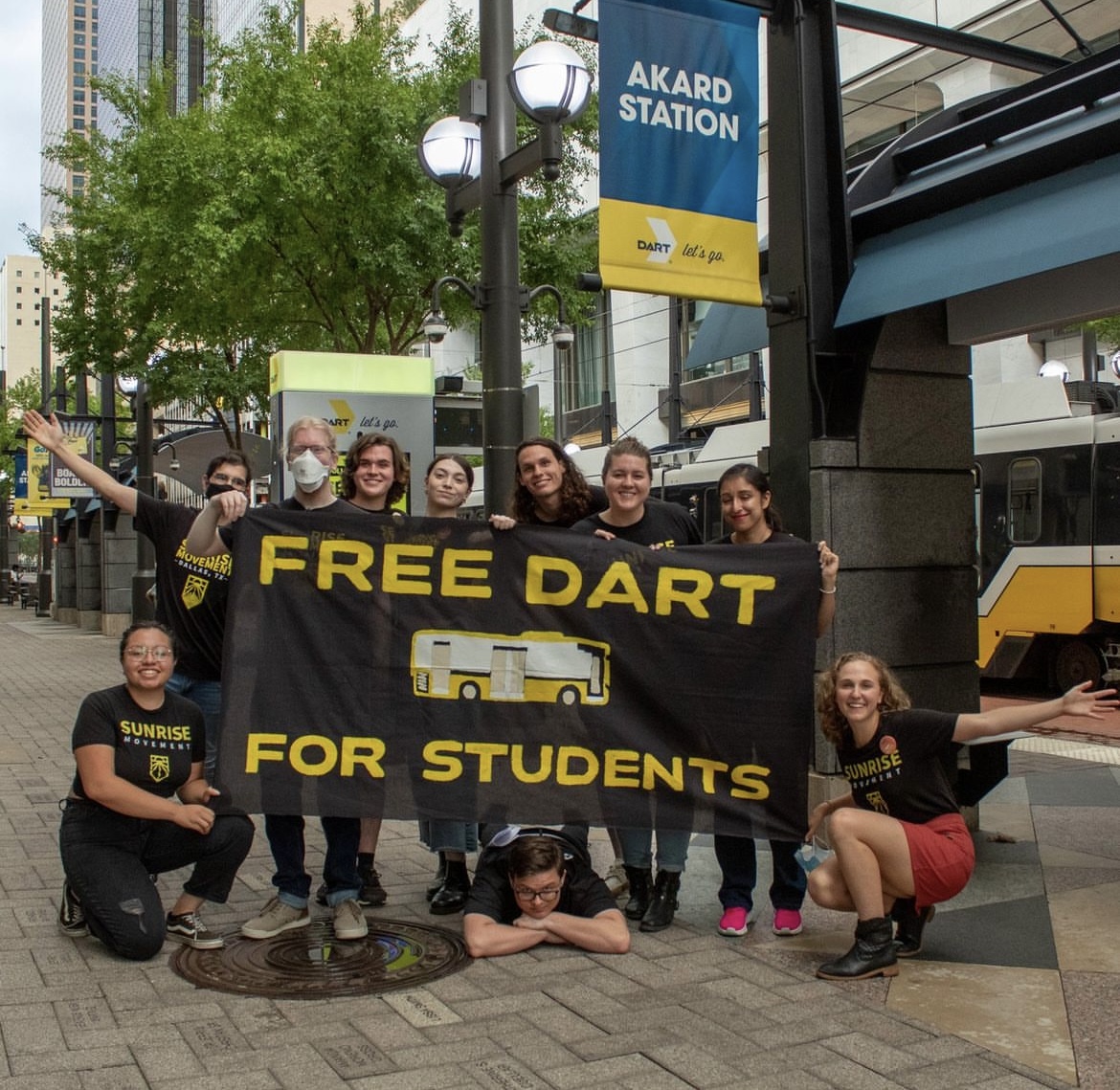 Sunrise Dallas members holding a Free DART for Students banner at Akard Station, Hope crouched at the right with arms wide