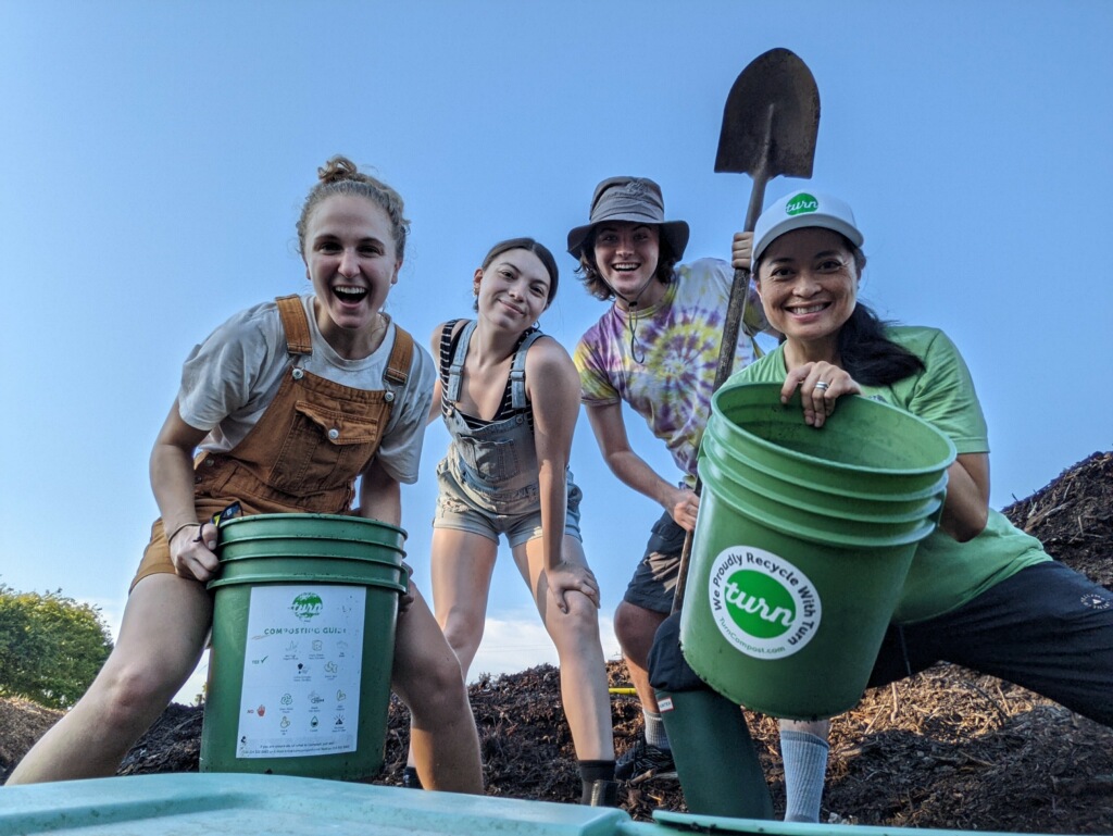 Hope in overalls with three friends grinning over green compost buckets and a shovel on a compost pile