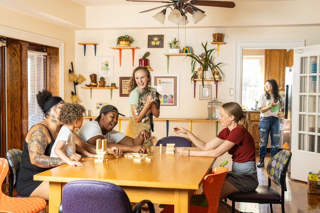 Housemates playing a block-stacking game at a wooden kitchen table while Hope laughs holding a cat, plants and shelves on the walls behind