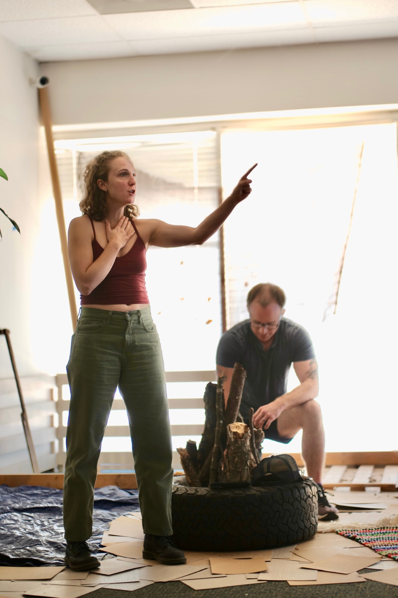 Hope standing with a hand on her chest and pointing across a rehearsal space, a collaborator arranging logs in a tire behind her