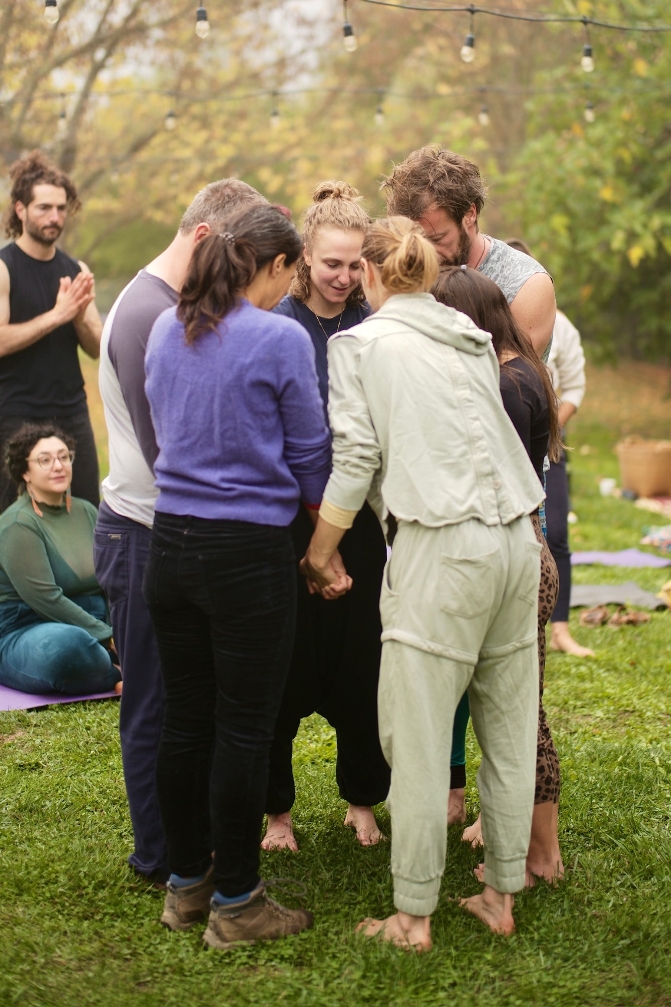 A small circle of people standing barefoot on grass holding hands with heads bowed, Hope at the centre, string lights overhead
