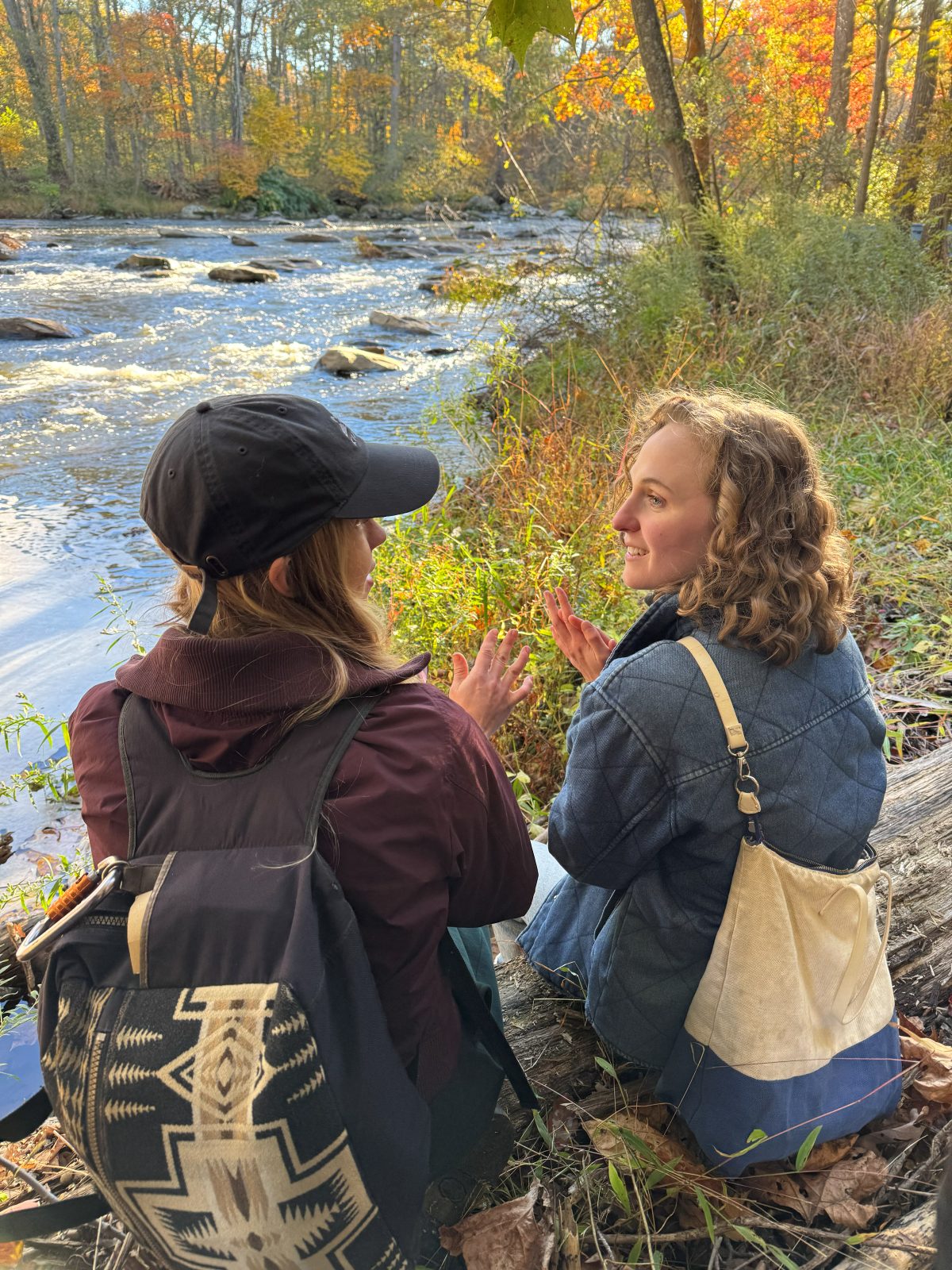 Hope and a friend sitting on a log by a river, deep in conversation among autumn trees