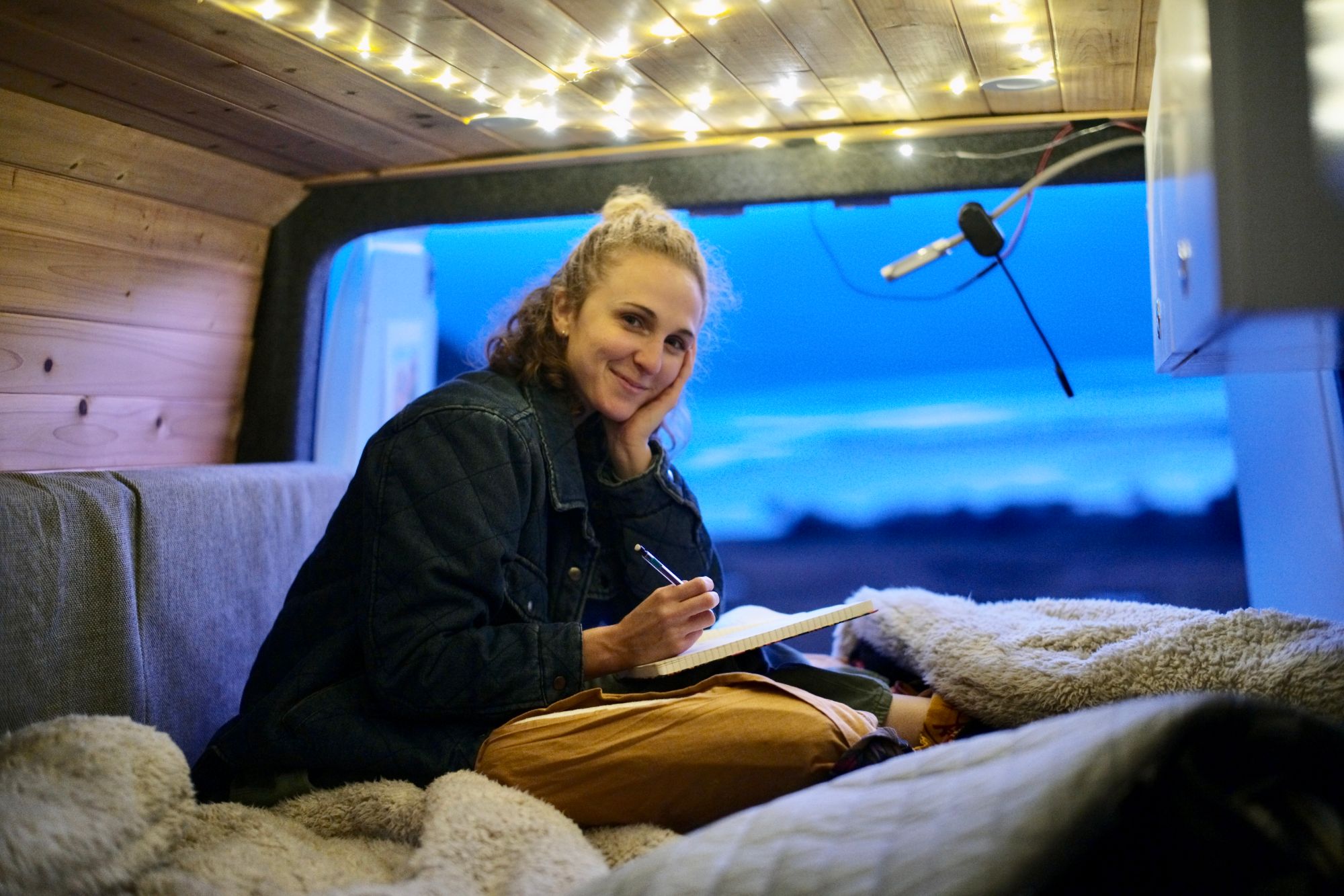Hope journaling on a cosy bed inside a wood-panelled camper van under string lights, the ocean at dusk through the window