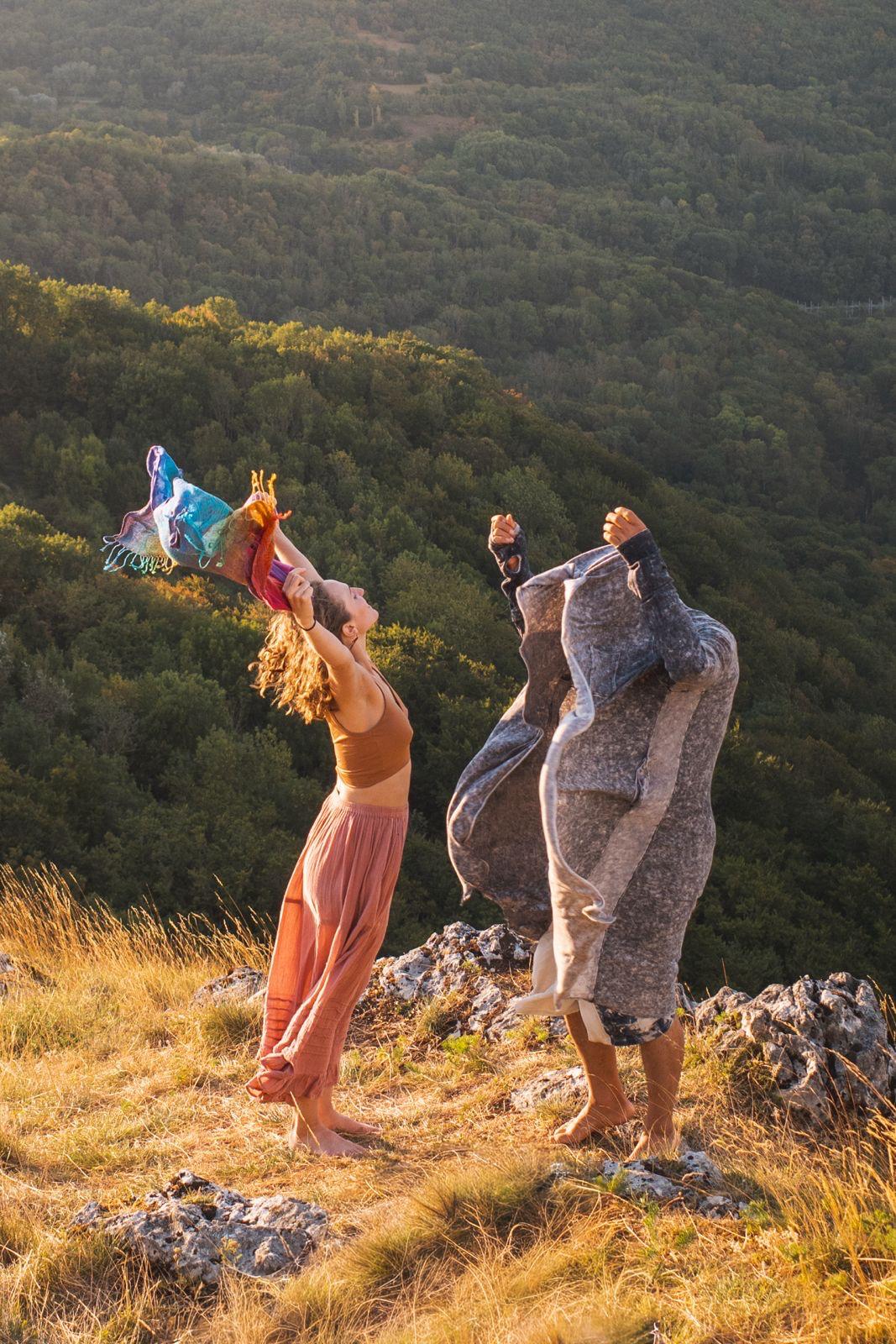 Hope dancing barefoot on a golden hillside at sunset, arms raised with a rainbow scarf, a friend beside her wrapped in a blanket