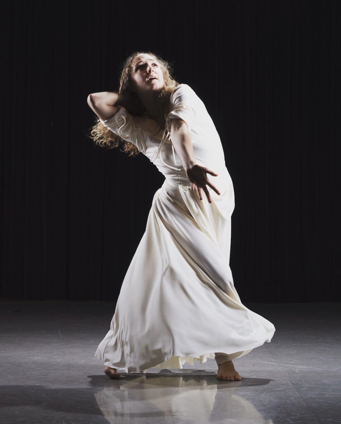 Hope dancing barefoot in a flowing white dress on a dark stage, arm reaching forward