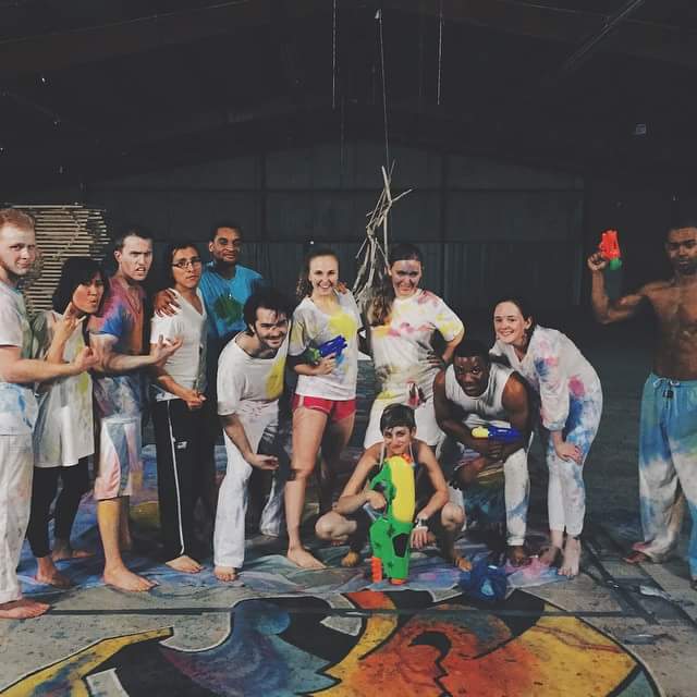 A cast of a dozen performers in paint-splattered white clothes posing with water guns on a painted floor