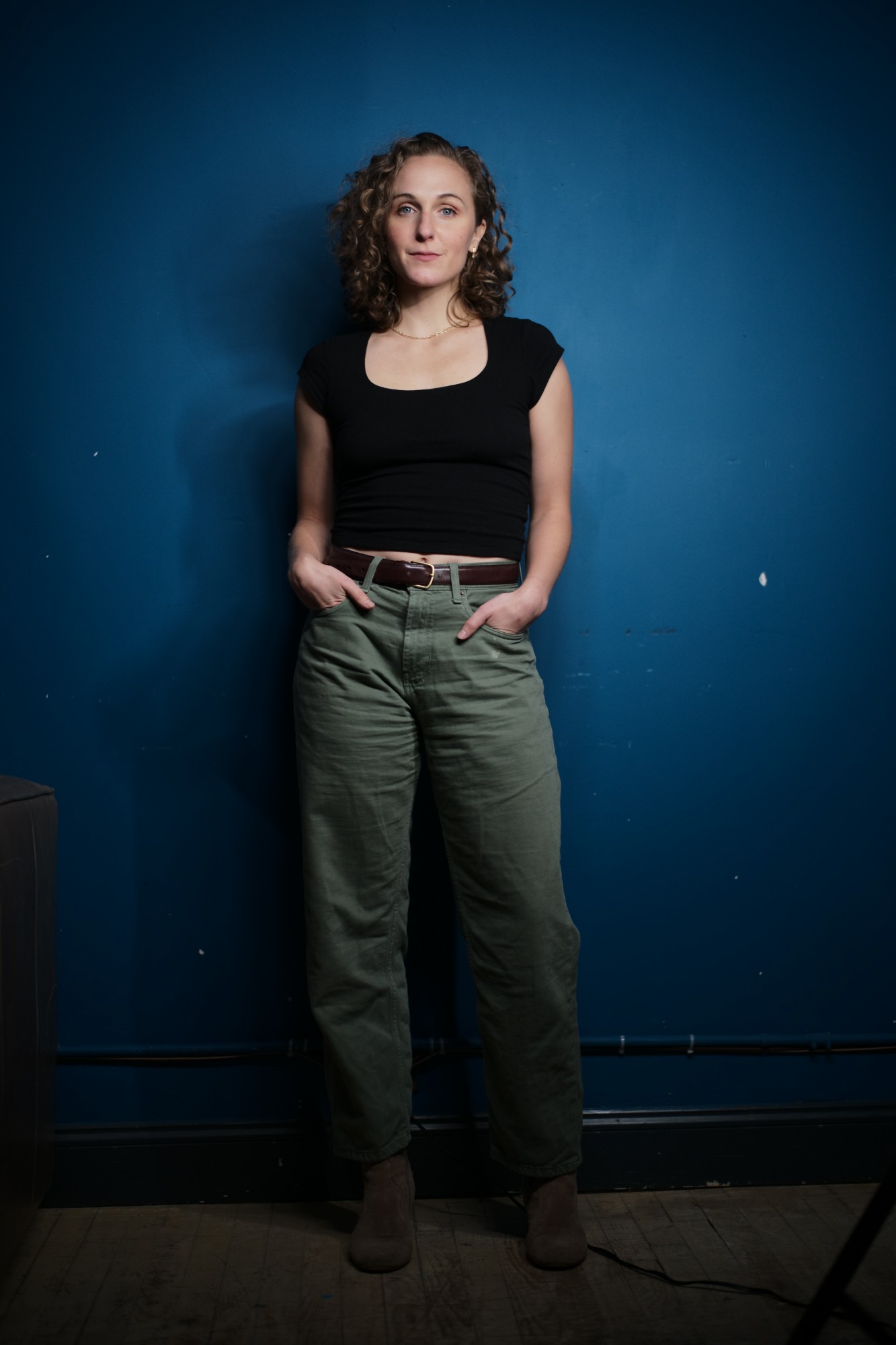 Hope standing against a deep blue wall in a black top and olive trousers, hands in pockets, looking at the camera