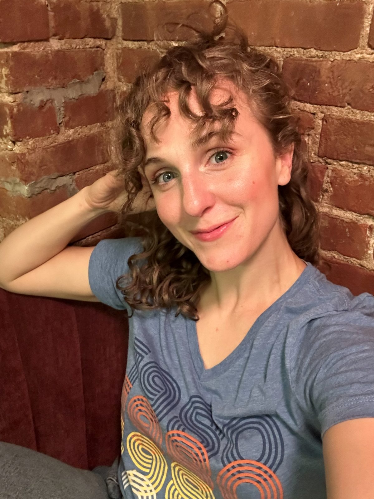 Hope leaning against a brick wall in a blue tee, hand in her curls, smiling softly